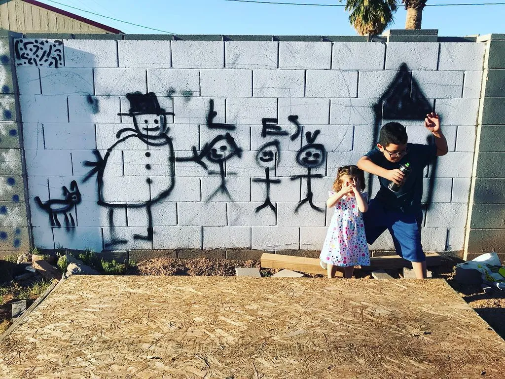 Black drawings of figures adorn a light gray cinder block wall, where an adult and a small child stand on a textured wooden surface.