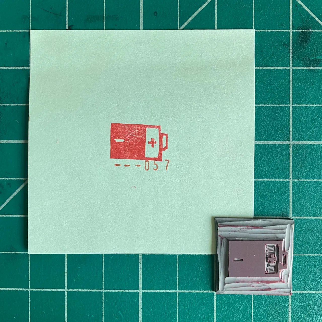 A light mint green square paper on a dark teal grid displays a red drawing of an ambulance and the number --057, positioned above two small, framed objects with red and white markings.