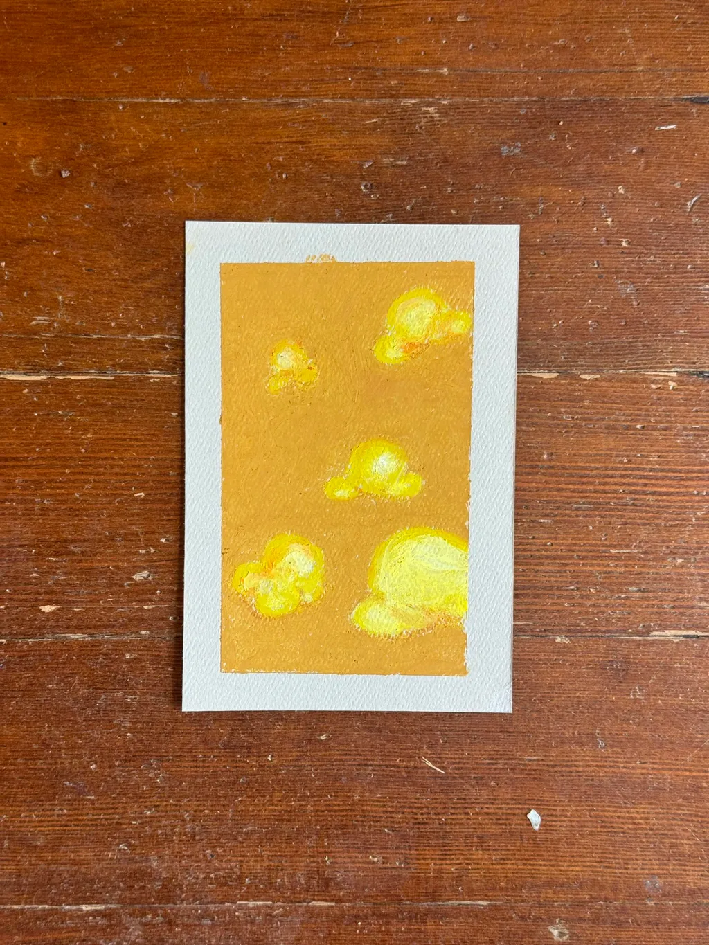 A vertical painting on a mustard yellow background features several scattered, irregular shapes of bright yellow and gold, painted with visible, soft brushstrokes.