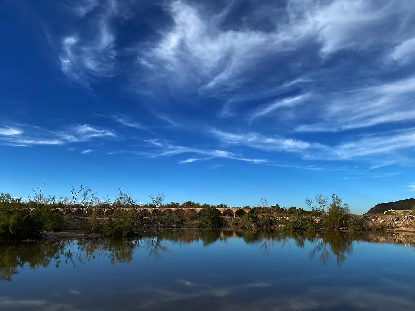 A wide landscape shows a calm, reflective blue water surface beneath a dramatic sky filled with streaky white and blue clouds, with a low line of dark green vegetation and structures along the horizon.