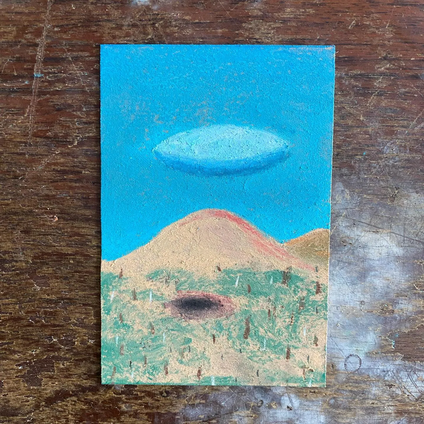Bright blue sky dominates the upper half, featuring a pale blue oval object above a tan landscape with a central reddish hill, sparse green marks, and a dark circular patch on the ground.