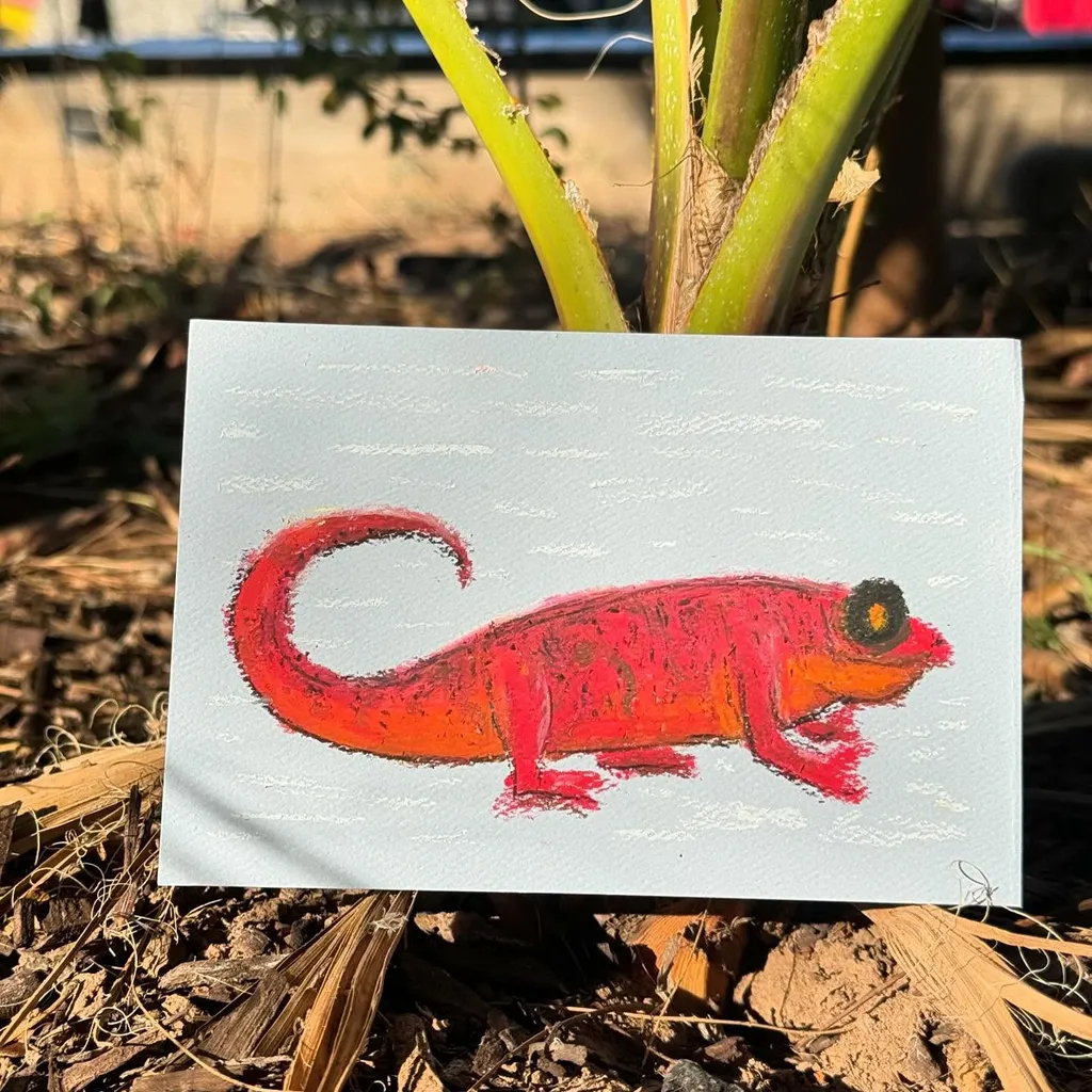 A bright red lizard drawing rests on a white card, positioned outdoors on dark mulch, with a vertical green stalk visible in the background.
