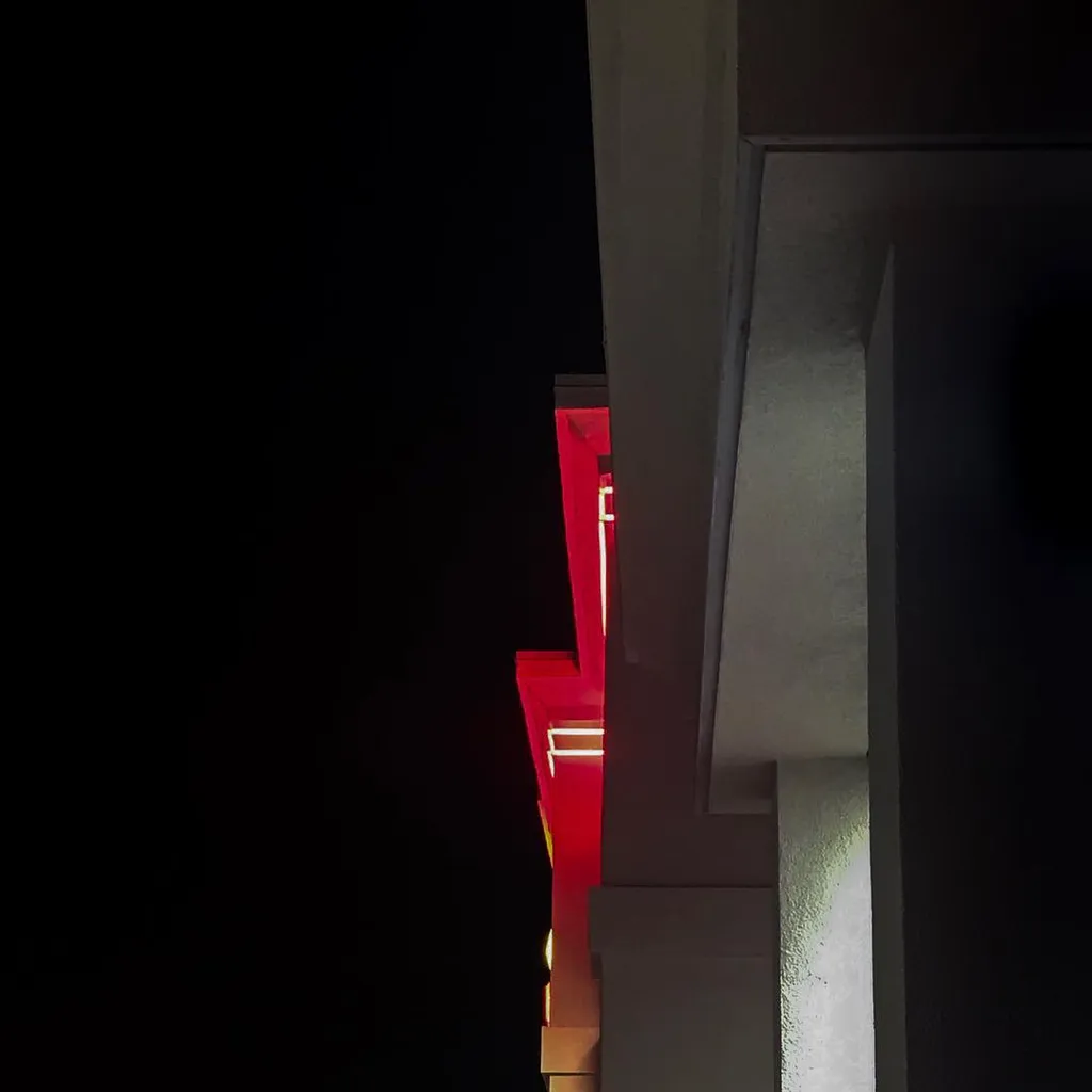 A vertical composition featuring smooth gray architectural planes on the right, contrasting with a deep black void on the left, where a recessed, glowing red structure is visible.