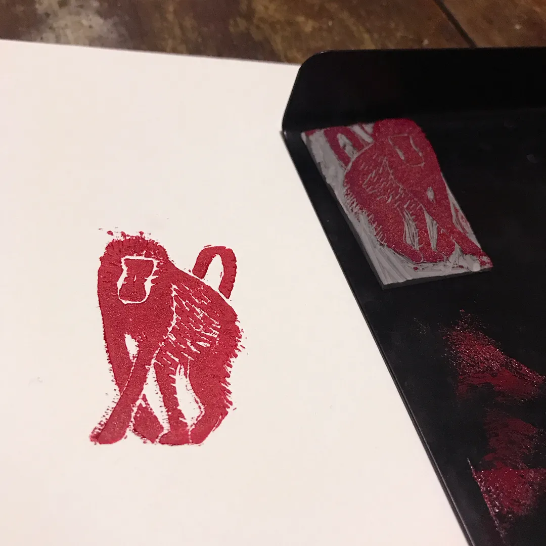 A red, abstract figure is painted on white paper next to a matching red figure displayed on a contrasting black panel, both rendered with rough, gestural strokes.