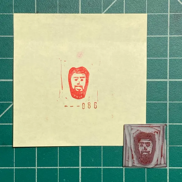 On a green grid, a cream paper displays a red stylized face drawing and the number --086, alongside two small, rectangular art pieces featuring red and white layered stripes and stylized faces.