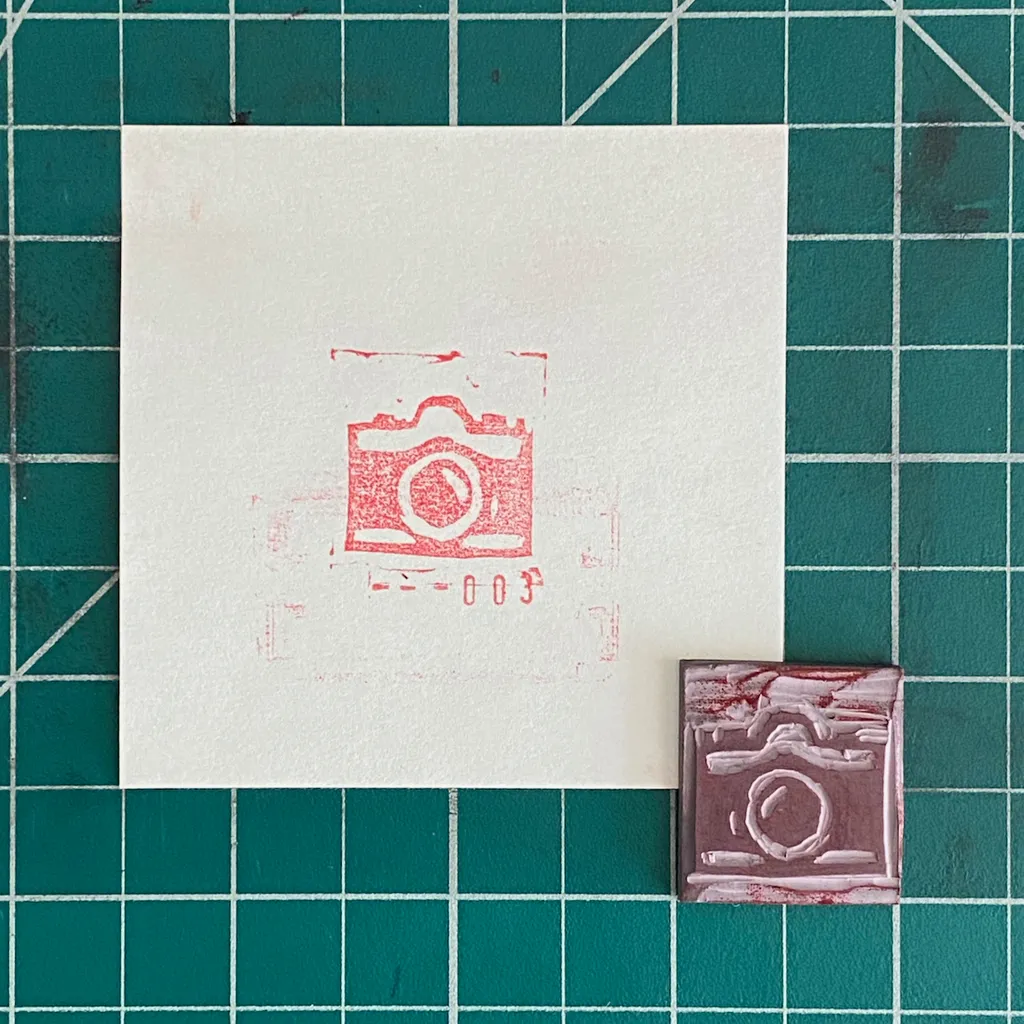 A white square paper with a red stamped camera image rests on a teal grid, alongside a small reddish-brown rectangular object featuring an embossed camera design.
