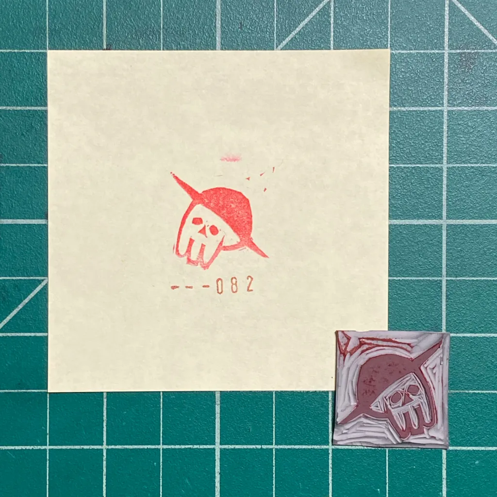 The composition features a cream paper on a teal grid, displaying a red drawing of a skull in a pointed hat and the number -- - 0 8 2, alongside a small, reddish-brown square with a matching skull design.