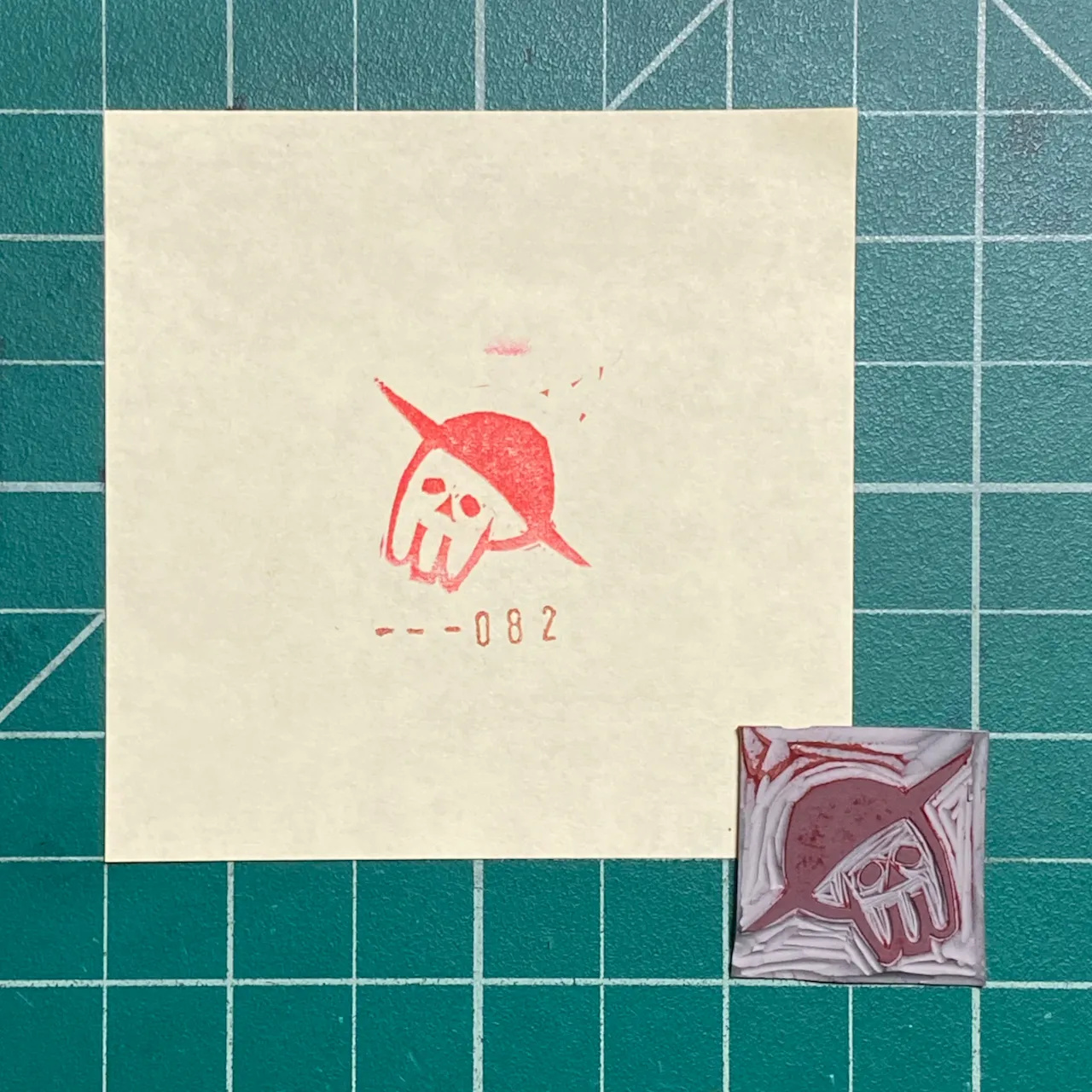 The composition features a cream paper on a teal grid, displaying a red drawing of a skull in a pointed hat and the number -- - 0 8 2, alongside a small, reddish-brown square with a matching skull design.