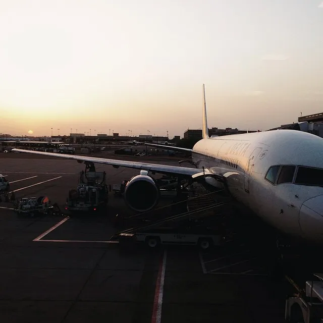 The white fuselage of a parked airplane occupies the right side, set against a dark tarmac and a soft gradient sky of pale yellow and orange at the horizon.