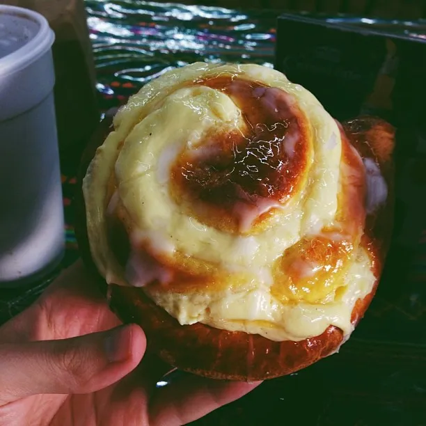 A hand supports a golden-brown baked roll covered in a thick, glossy topping of melted yellow and white cheese, which drips down the sides.