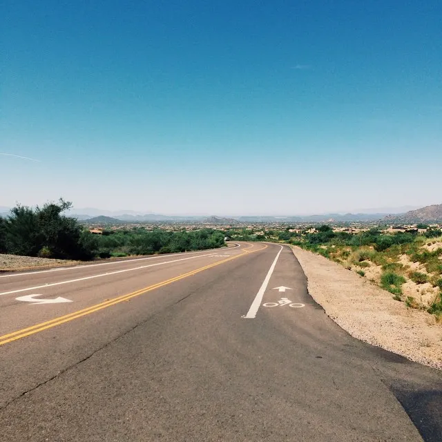 A paved road curves through an arid landscape under a bright blue sky, showing yellow dividing lines and white painted bicycle symbols on the asphalt.