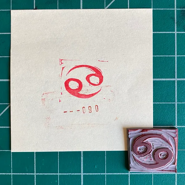 A cream paper on a green grid displays a red circular swirl and the numbers 0-090, positioned above a small square piece featuring layered pink and maroon swirls.