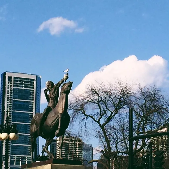 A bronze sculpture of a man riding a rearing horse dominates the foreground against a bright blue sky filled with white clouds, flanked by modern buildings and dark trees.