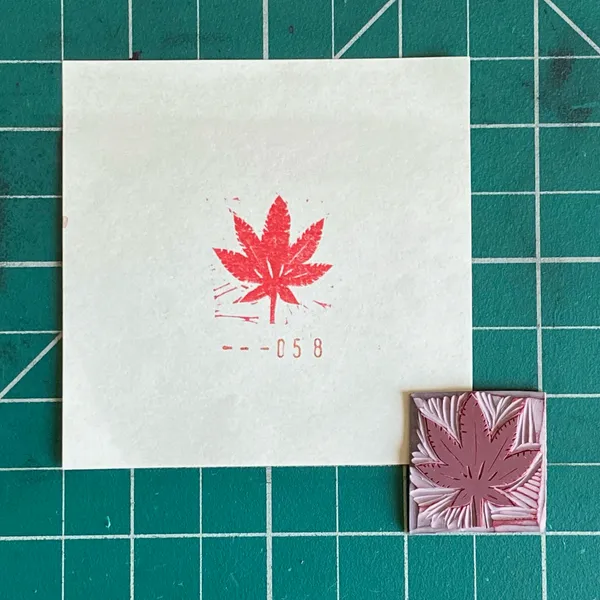 A white card displaying a red maple leaf and the numbers 058 sits on a green grid background next to a small, carved pink piece featuring a textured maple leaf.