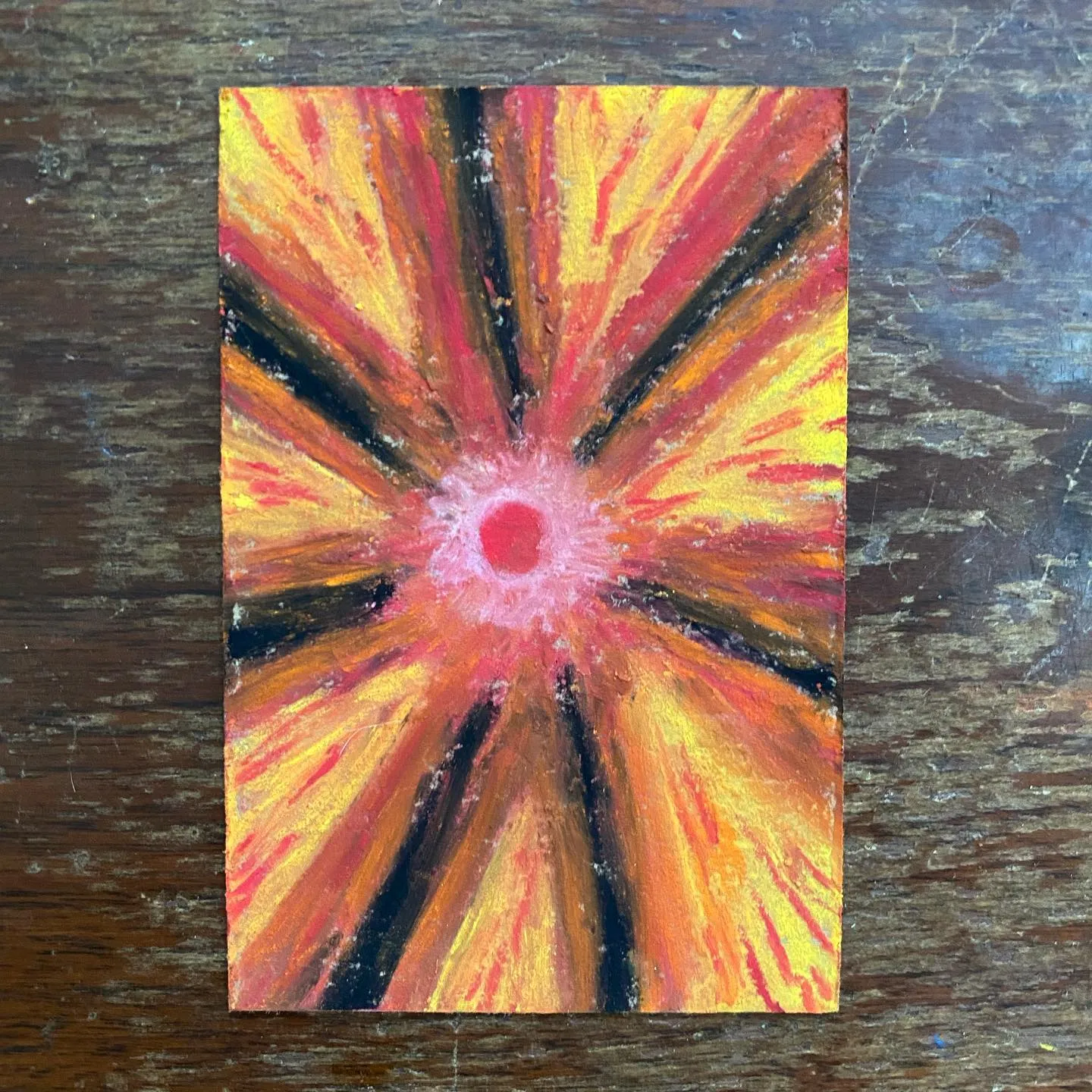 A square painting features a vibrant, radial starburst pattern of yellow, orange, and red paint radiating from a small pink center, accented by black lines, set against a dark wooden background.