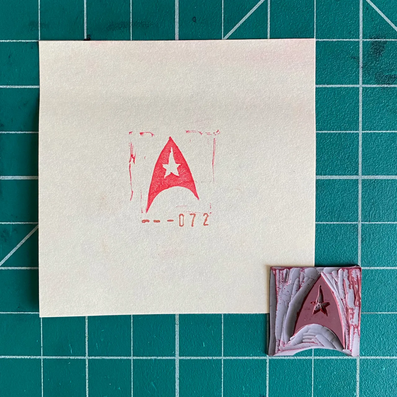 A cream sheet of paper on a green grid displays red pencil markings for a star-adorned pennant, next to a small, textured piece featuring muted pinks, grays, and a red star.