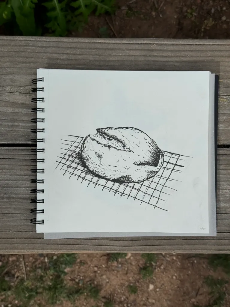 A graphite sketch of a loaf of bread, shaded with visible marks, rests on a pattern of diagonal hatching lines on a page in a spiral notebook.
