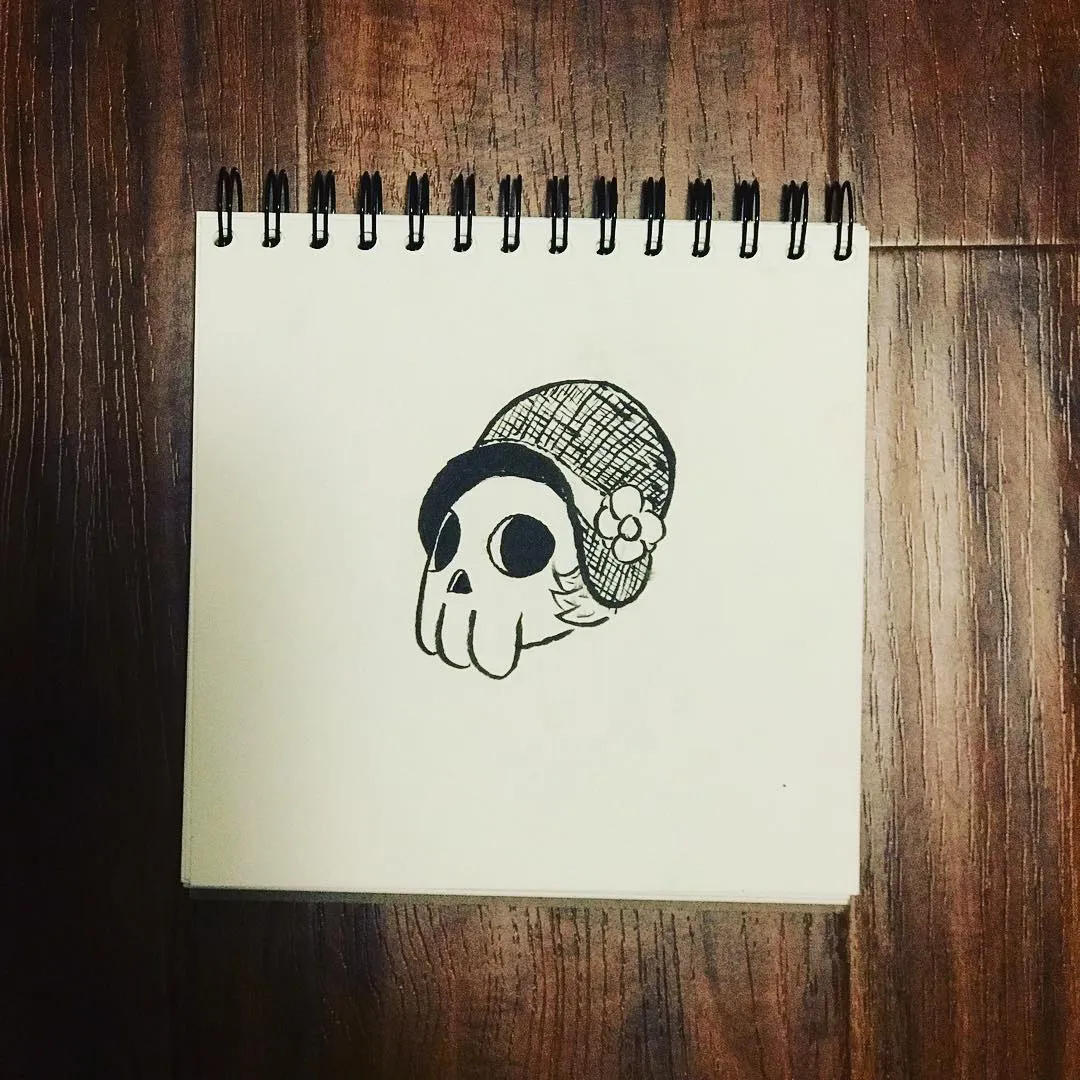 A black ink drawing of a stylized skull wearing a patterned cap and a small flower, drawn on white paper and placed on a dark wooden surface.