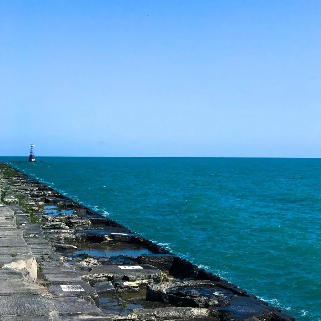 Rough, wet gray stones form a jetty stretching into deep turquoise water beneath a vast, clear blue sky, marked by a distant red beacon.