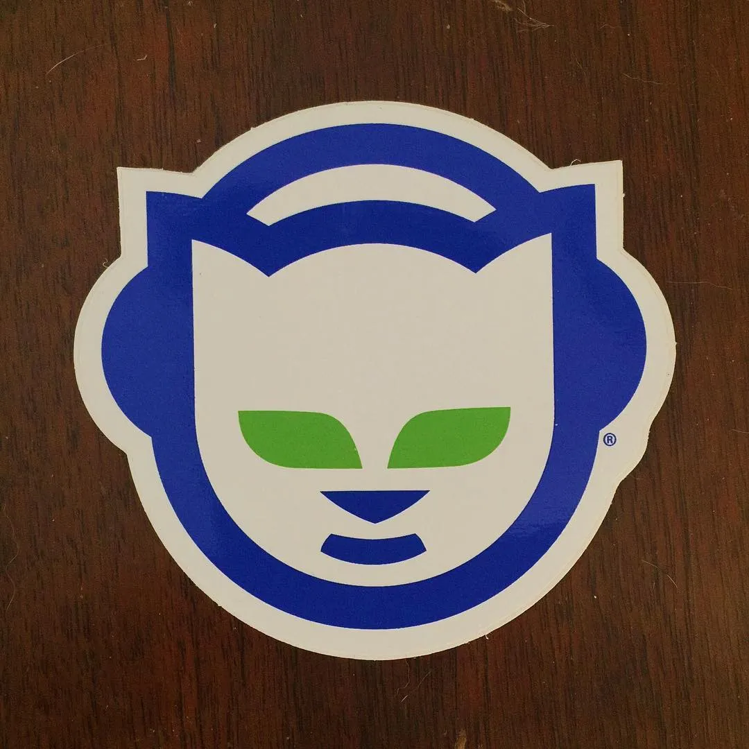 A stylized cartoon mask featuring a white face, blue trim, and large blue ear shapes, with bright green eyes, set against a dark brown wood grain background.