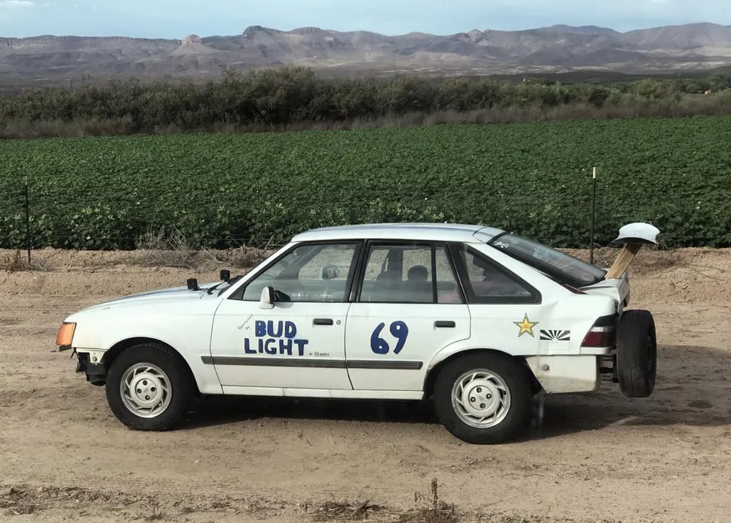 A white sedan with blue and black decals featuring BUD LIGHT and 69 sits on dirt ground, backed by a field of green crops and distant arid mountains.
