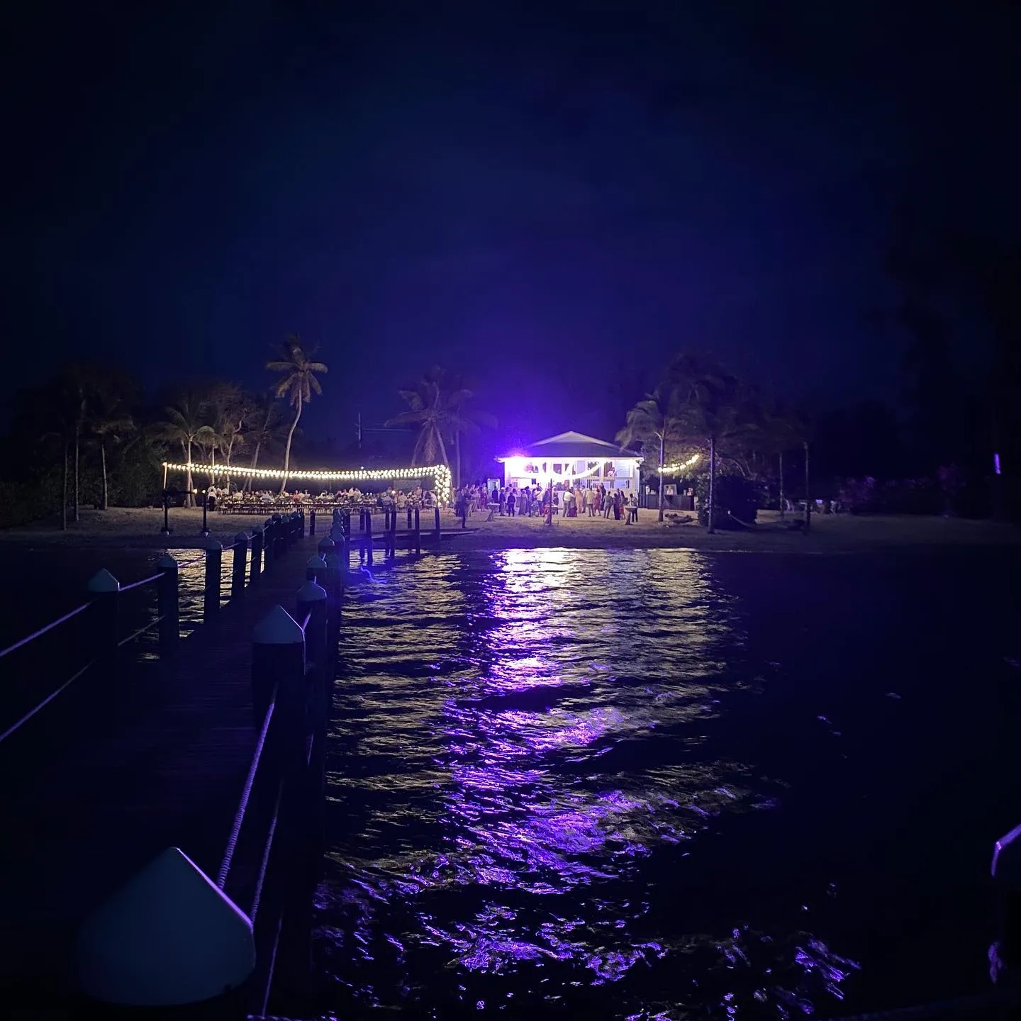 Dark water reflects intense purple light in the foreground, leading to a pier that crosses toward a brightly lit beach gathering under a dark night sky.