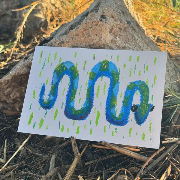 A stylized, wavy blue and green creature is drawn on white paper, featuring connected serpentine shapes and bright green accents, resting on rough rocks and dry earth.