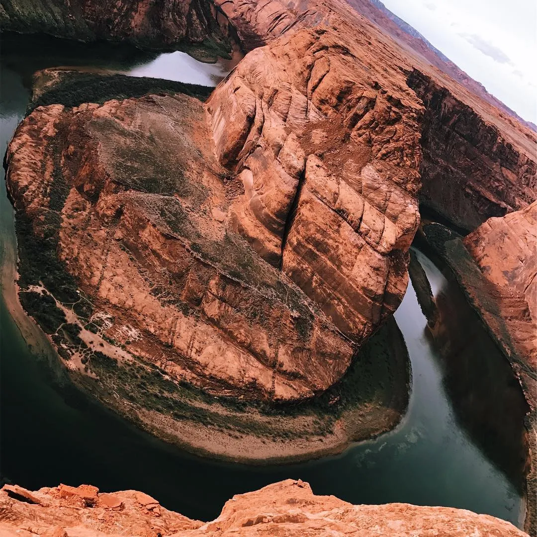 Aerial view of a deep gorge where stratified reddish-brown rock cliffs meet dark teal water, showing dramatic erosion and layered textures.