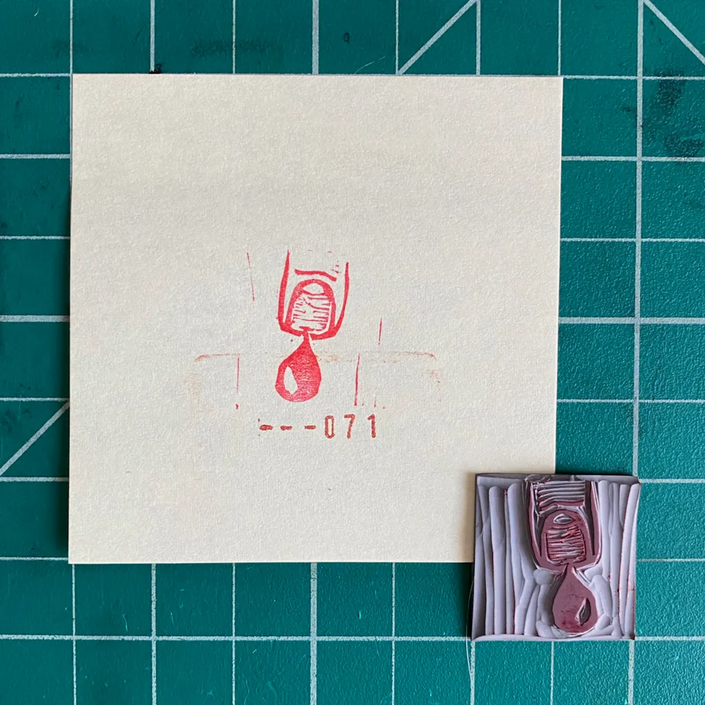 The composition features a cream square paper with a red ink drawing of a stylized figure and the number --071, next to a small, ridged pink rectangular object that also displays a matching stylized figure.