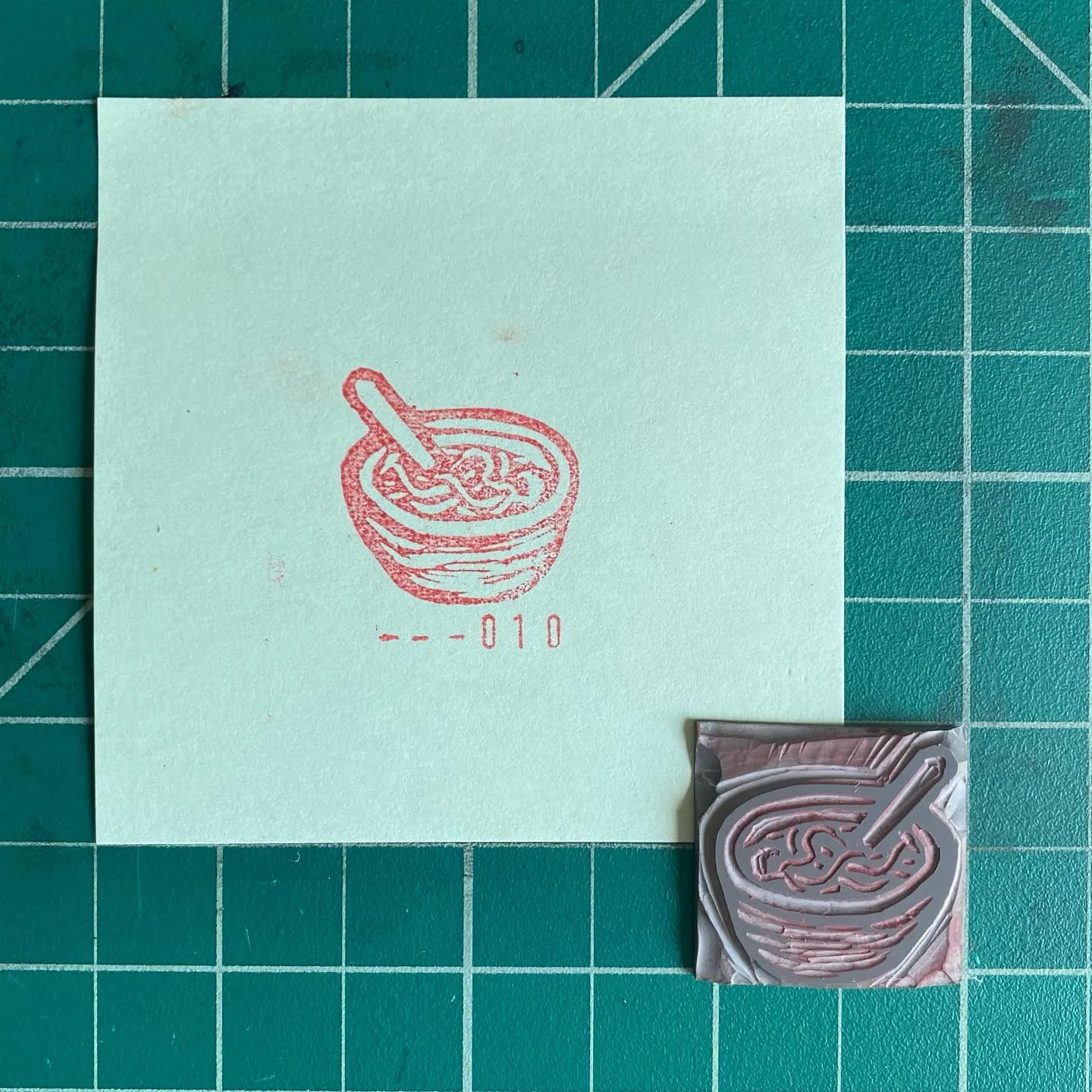 A pale cream square paper on a teal grid background displays a red drawing of a noodle bowl and chopsticks, marked with -010, next to a small, glossy square featuring the same red drawing.