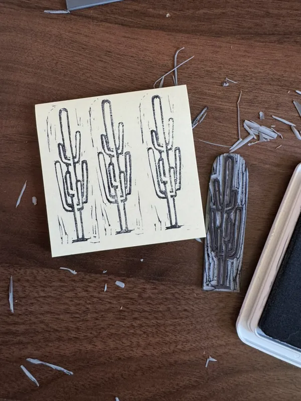 A stack of three drawings of cacti on cream paper rests on a dark brown wooden surface scattered with shavings, alongside a vertical textured drawing and a rough, gray wooden object.