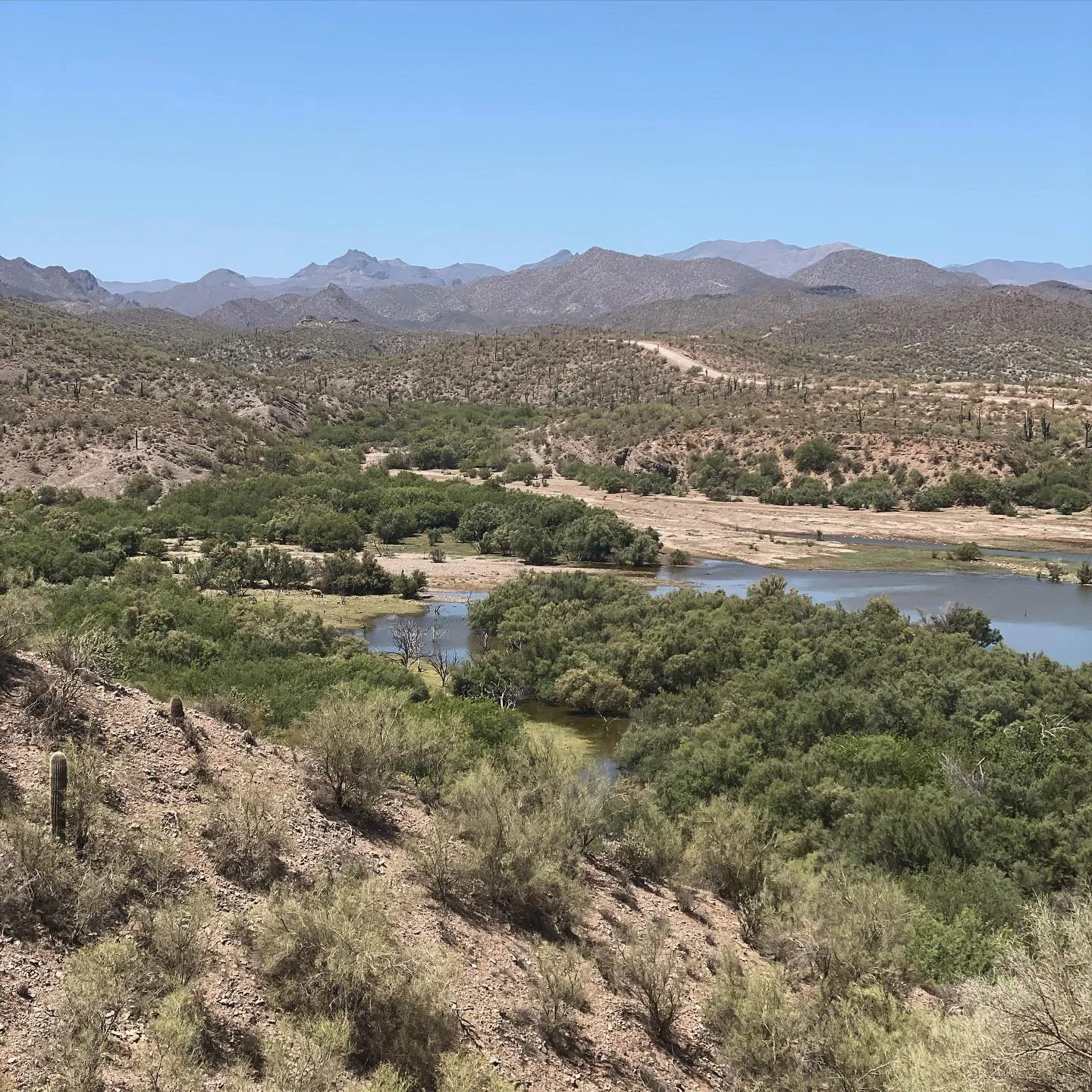 Dry, scrubby foreground slopes toward a calm body of water surrounded by dense green foliage, with rolling, arid hills and hazy blue mountains under a clear sky.