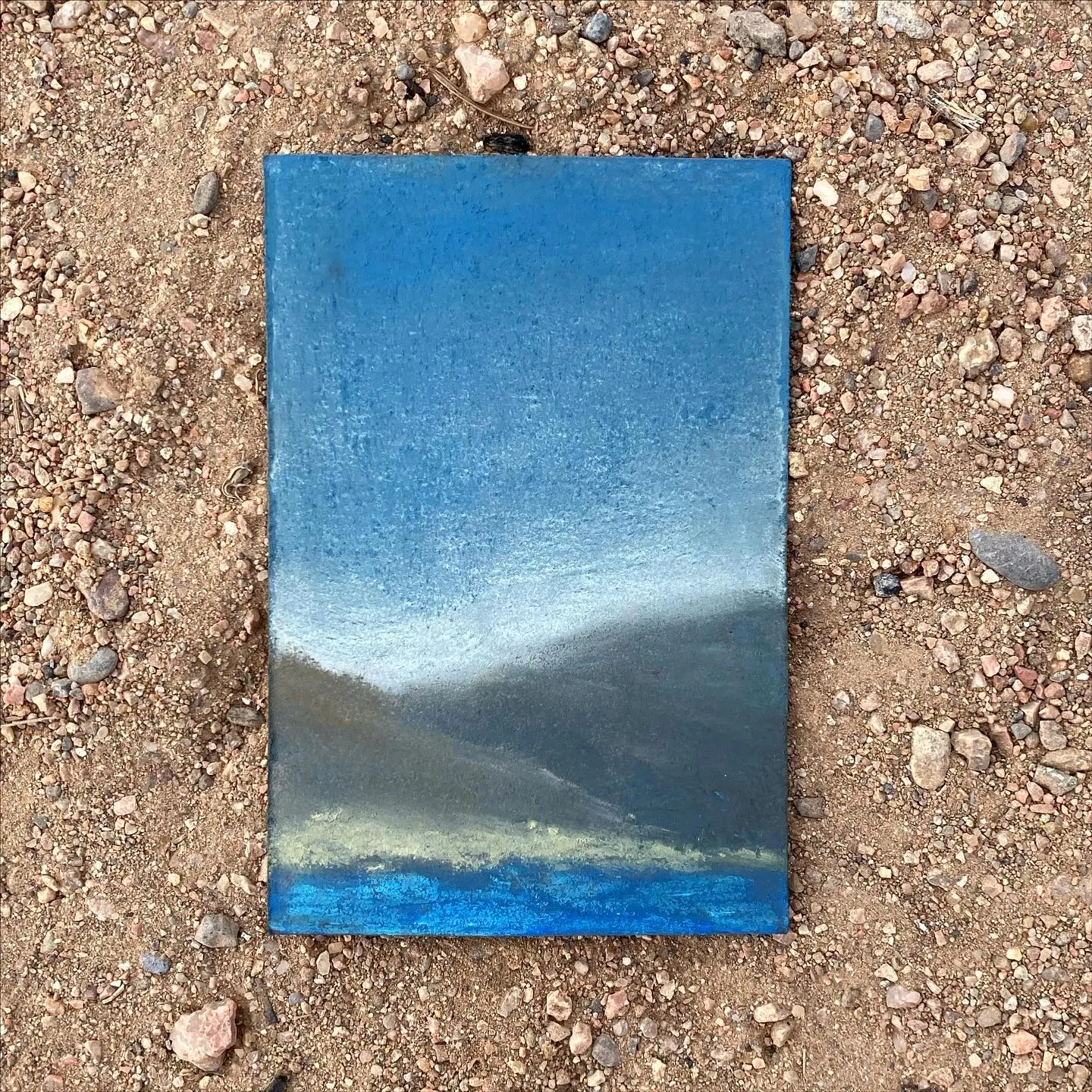 A rectangular painting depicting a horizontal landscape composition with pale blue sky, dark gray distant land, and deep blue water, resting on gravel.
