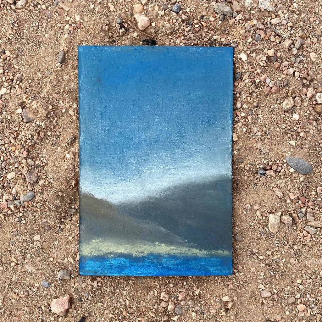 A rectangular painting depicting a horizontal landscape composition with pale blue sky, dark gray distant land, and deep blue water, resting on gravel.