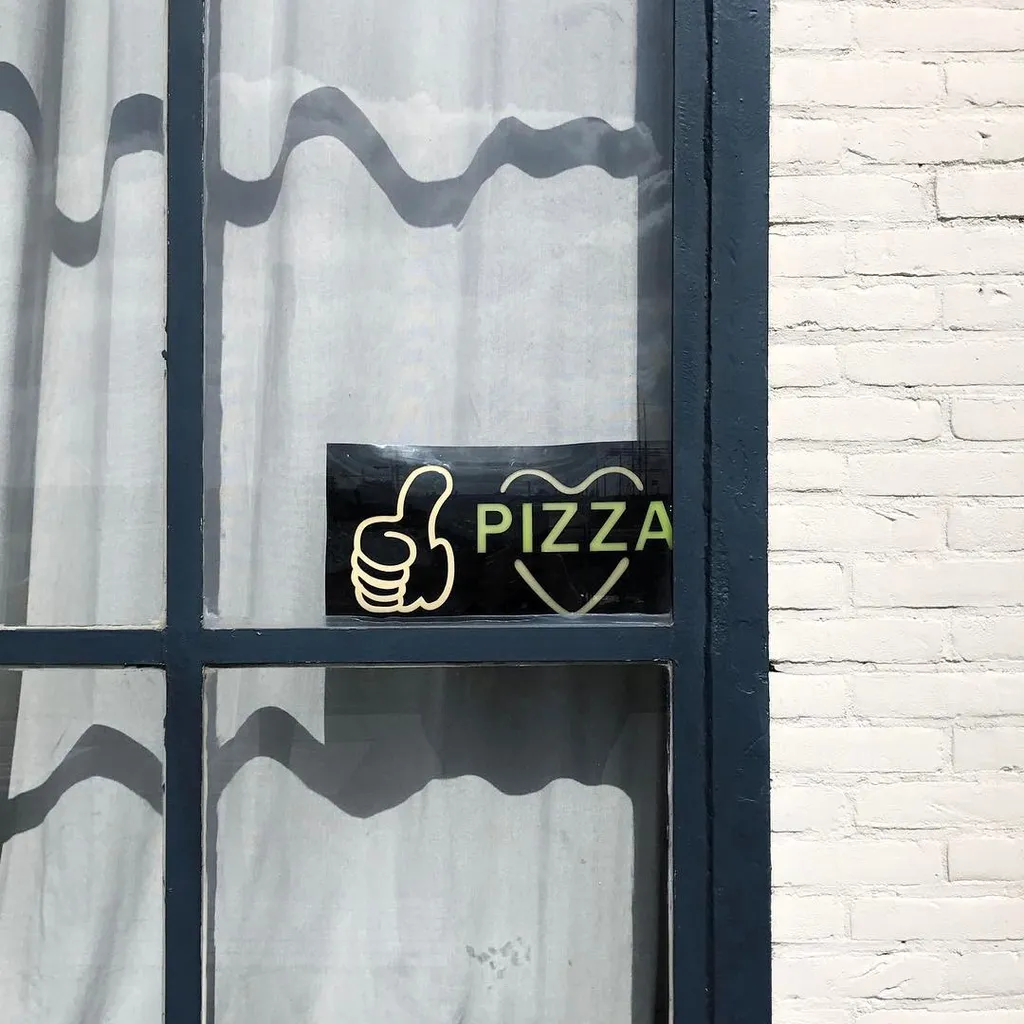 On a window pane set in a dark frame, a yellow graphic of a hand and the word PIZZA in a heart is visible against the backdrop of wavy shadows and white brick.