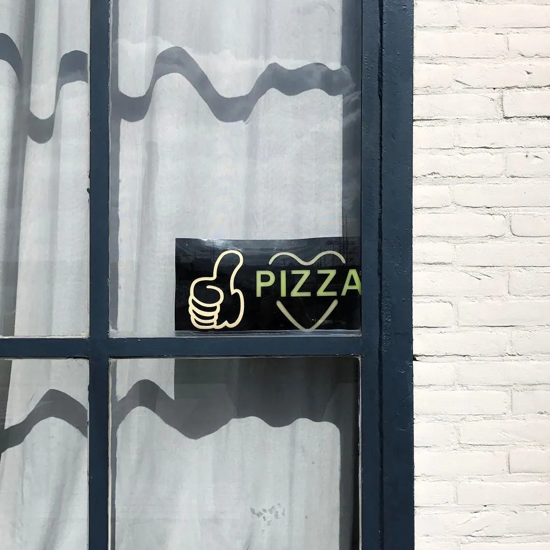 On a window pane set in a dark frame, a yellow graphic of a hand and the word PIZZA in a heart is visible against the backdrop of wavy shadows and white brick.