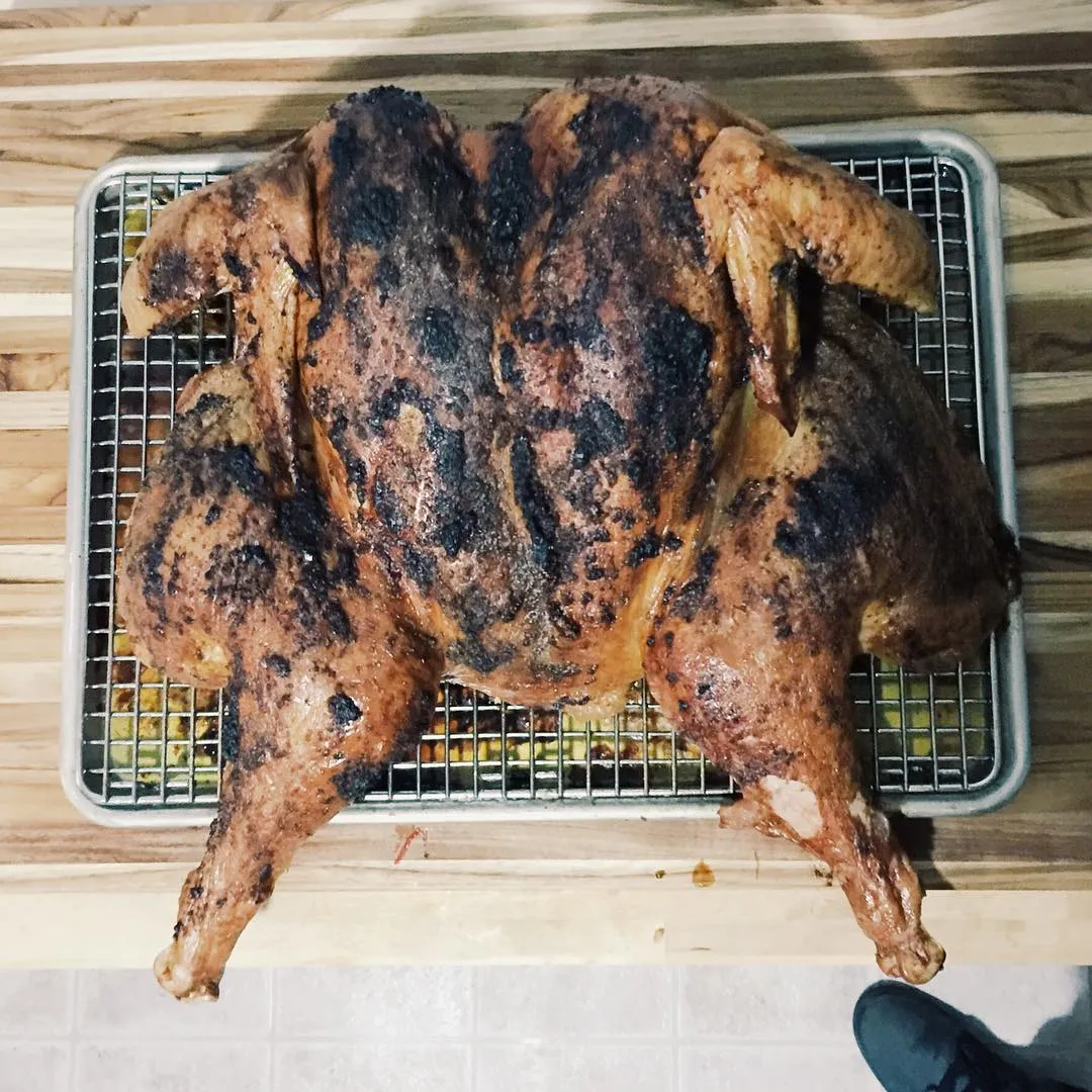 A whole roasted chicken with dark, crispy skin rests on a metal wire rack on a wooden surface.
