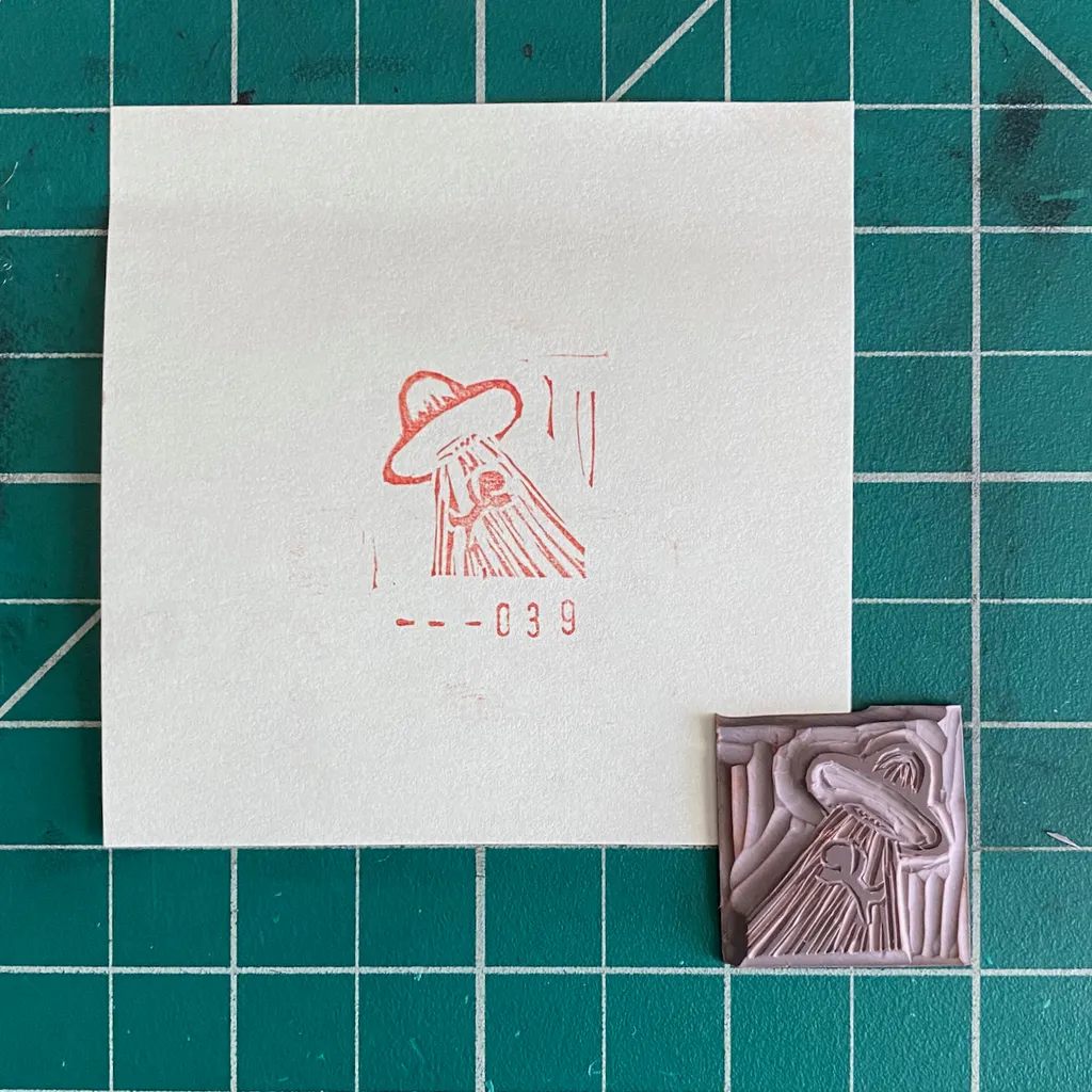 A white square displays a red drawing of a wide-brimmed figure and the number 039, positioned above a small, carved pinkish-brown square showing a textured version of the figure, all against a teal grid.
