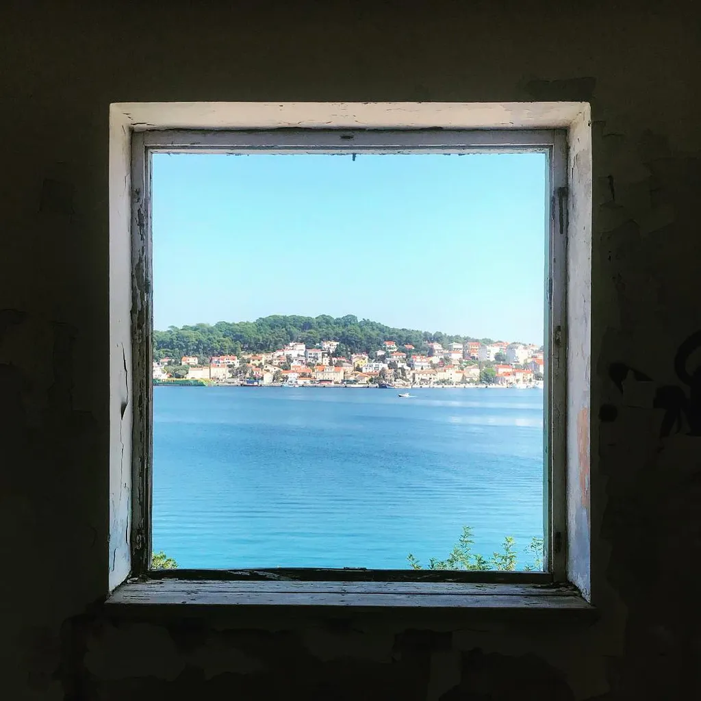 A view through a weathered, dark window frame shows a bright blue sky meeting calm blue water, with a densely built shoreline of terracotta buildings and green trees in the distance.