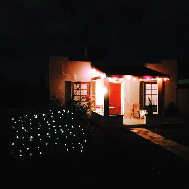 The composition features a warmly lit, single-story building at night, dominated by a red door and windows, contrasted by the deep black sky and a cluster of bright white lights in the foreground.