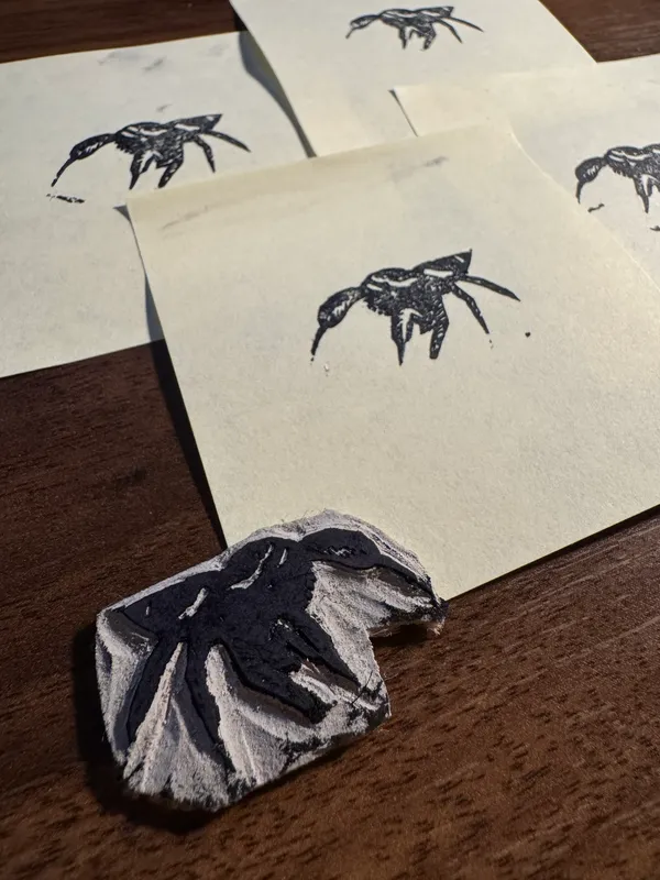 Black ink drawings of stylized spiders are scattered across overlapping sheets of off-white paper on a dark wooden surface, with a small, textured black and white object in the foreground.
