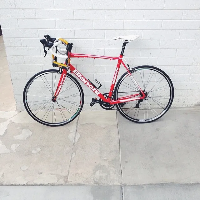 A bright red road bicycle rests on a light concrete floor against a white painted brick wall, featuring black tires and components.