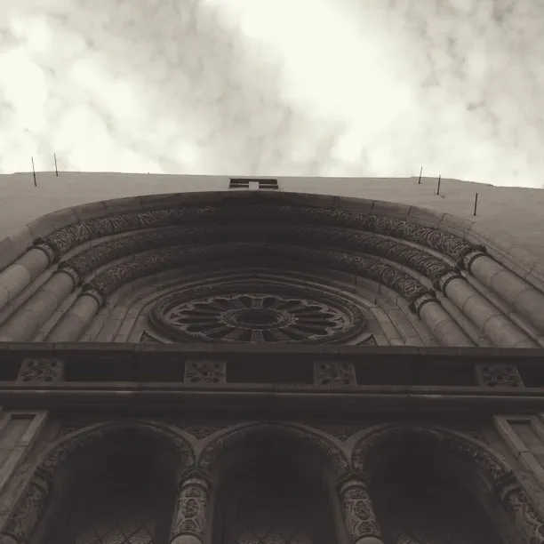 A low-angle view of a dark, textured stone facade featuring a large semicircular arch and central rose window, set against a bright, cloudy sky.