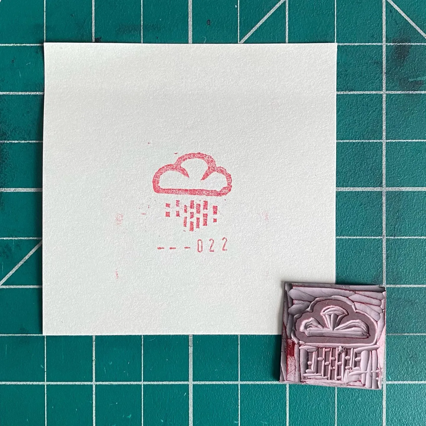 A white square on a teal grid displays a red cloud drawing and the numbers --022, next to a small, pale pink square featuring raised geometric patterns and a cloud motif.