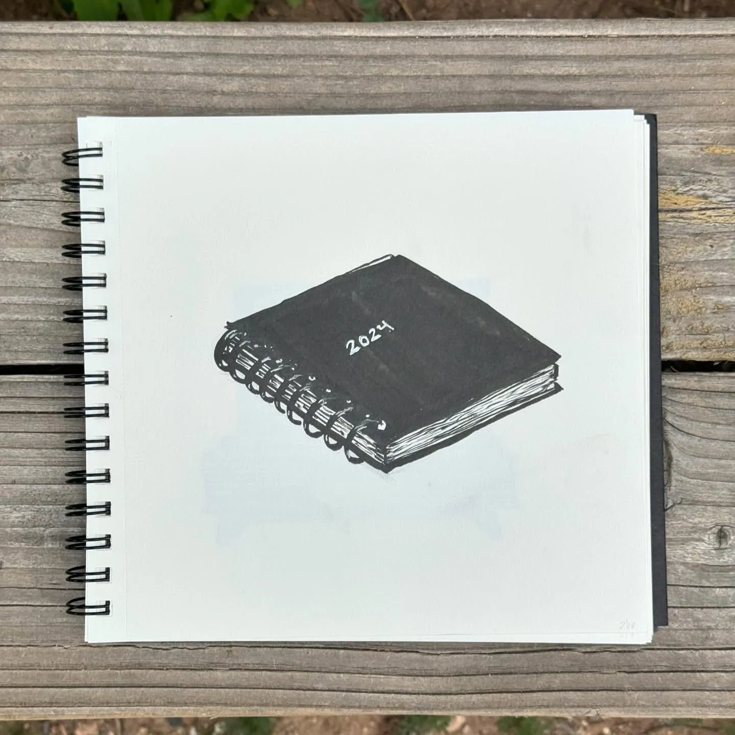 A drawing of a dark spiral notebook marked 2024, centered on white paper that rests on a textured brown wooden surface.