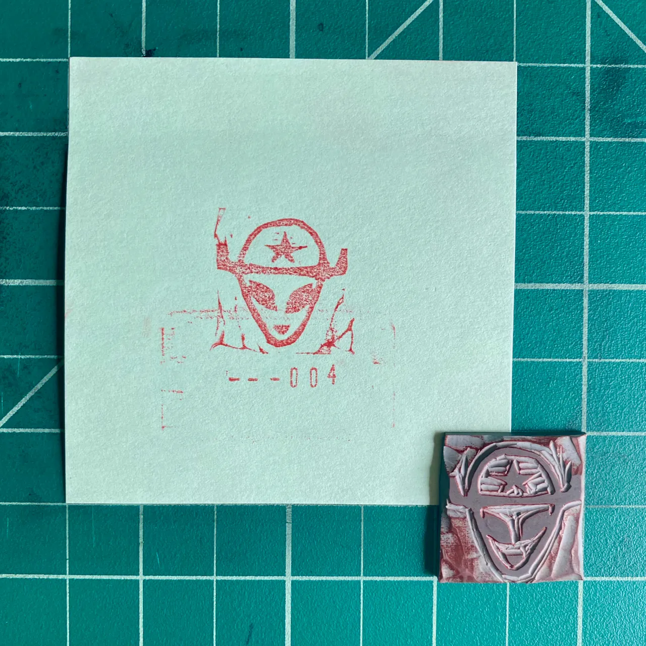 On a teal grid, a white paper displays a red printed mask design and the number —004, next to a small, reddish-brown square object featuring an embossed mask.