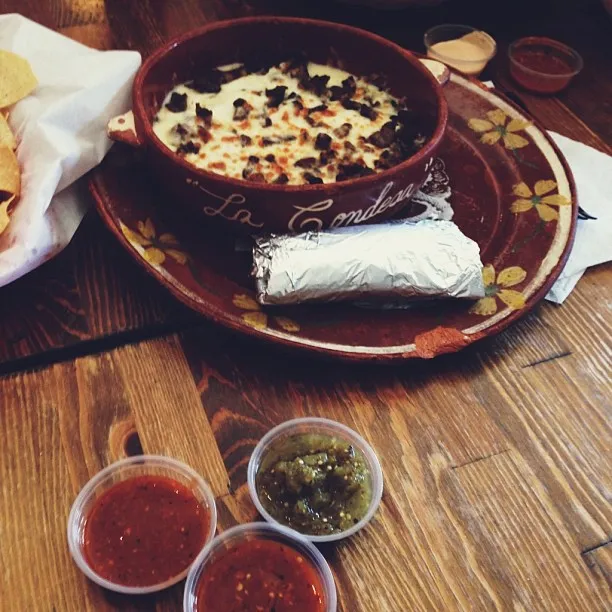 Overhead view of a wooden table featuring a central, dark bowl with white baked food, resting on a patterned plate, flanked by small bowls of red, green, and brown dips.