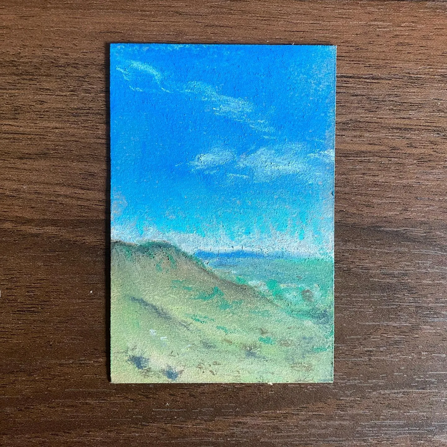 The artwork depicts a landscape with a vibrant blue sky showing wispy white clouds, meeting a horizontal band of muted sage green hills and distant ground.