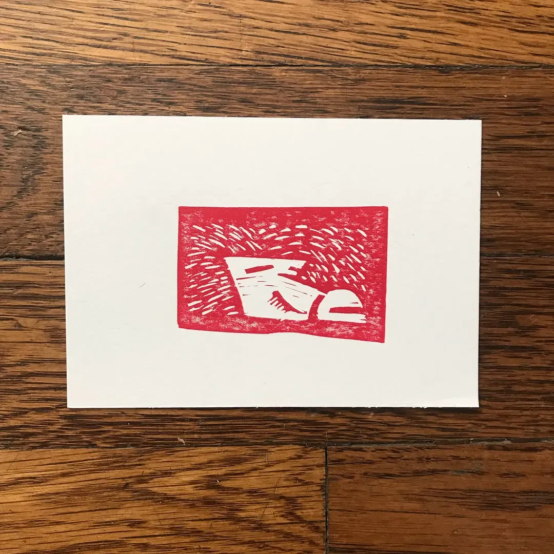 A distressed red square graphic with abstract, angular shapes is centered on a white card resting on a brown wooden surface.