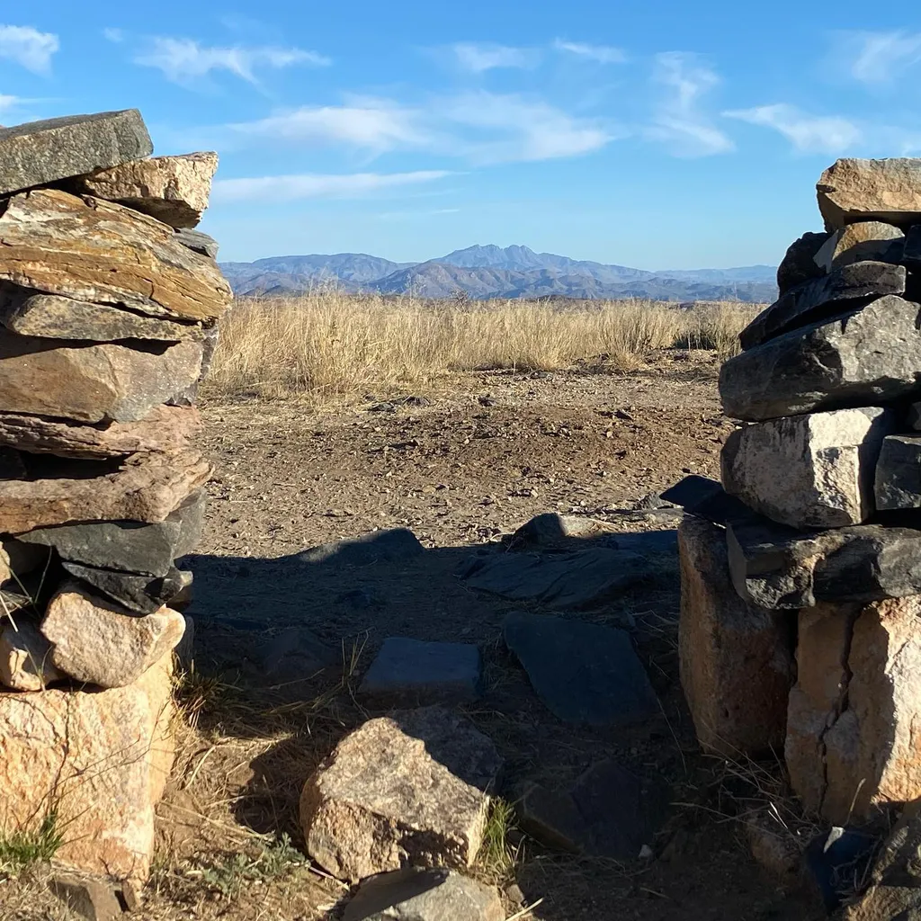 Rough stacked stones frame a dry, tan landscape that stretches across the midground toward a distant, hazy mountain range beneath a bright blue sky.